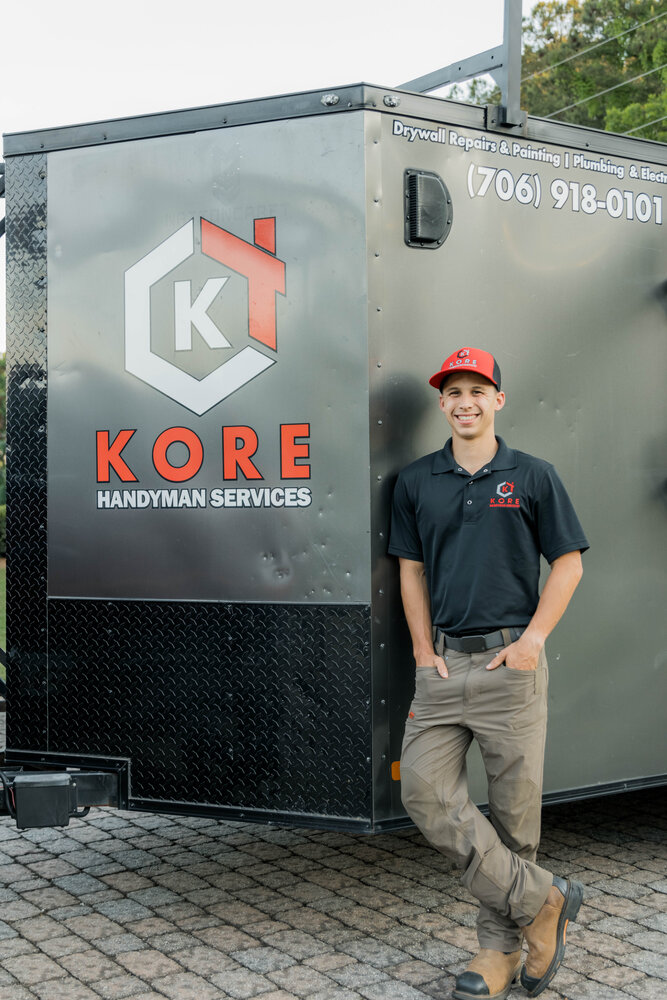 Kade Bloom's smiling employee Nathaniel standing in a white shirt with the red and black KORE logo, with trees and a stone wall behind him.