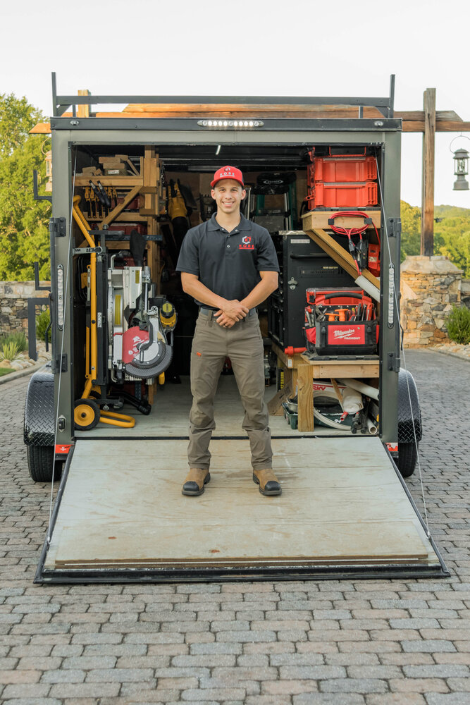 Kade Bloom's black and red KORE workshop trailier has the door open and laying as a ramp on the ground, and he's standing in the middle of the ramp with arms crossed and a big smile on his face