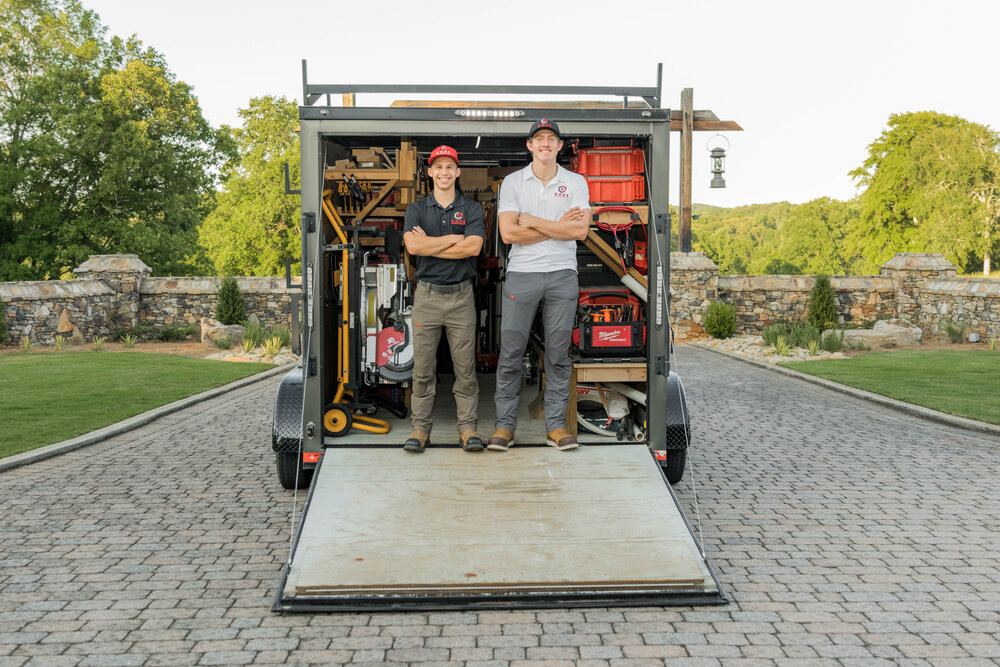 Kade Bloom's black and red KORE workshop trailier has the door open and laying as a ramp on the ground, and he and his employee Nathaniel are standing together at the door.
