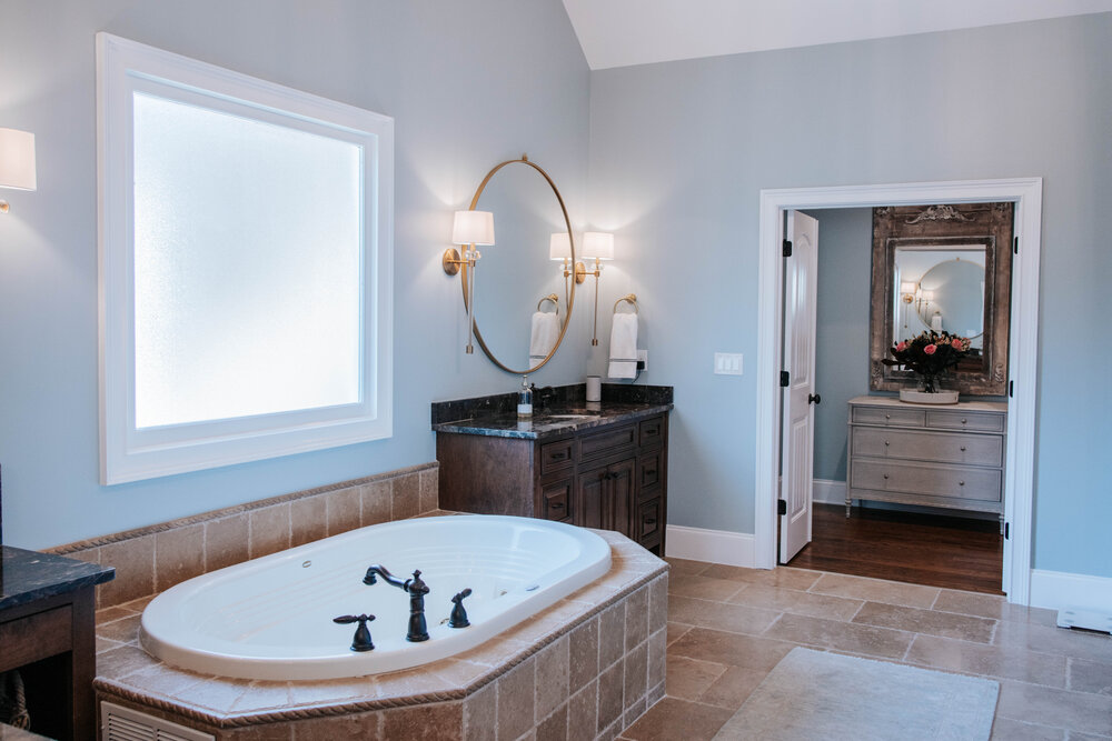 A relaxing bathroom with large spa tub surrounded by tile work, dark wood sink cabinet, large round mirror with light sconces on both sides.