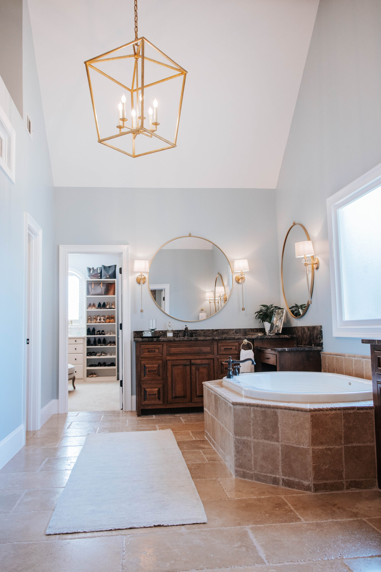 A large, high-ceiling bathroom with a spacious tile-covered spa tub, tile flooring, a large dual-sink cabinet with large round dual mirrors above it.