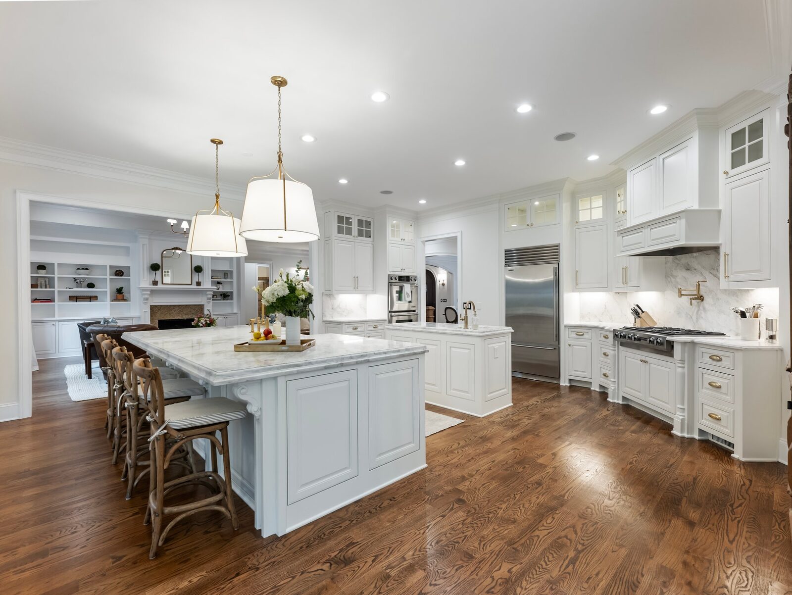 A spacious open-plan kitchen with a large breakfast island with hanging ceiling lamps above it, a smaller white marble-top work island to the side, and a stainless steel refrigerator and other white cabinetry along the wall.