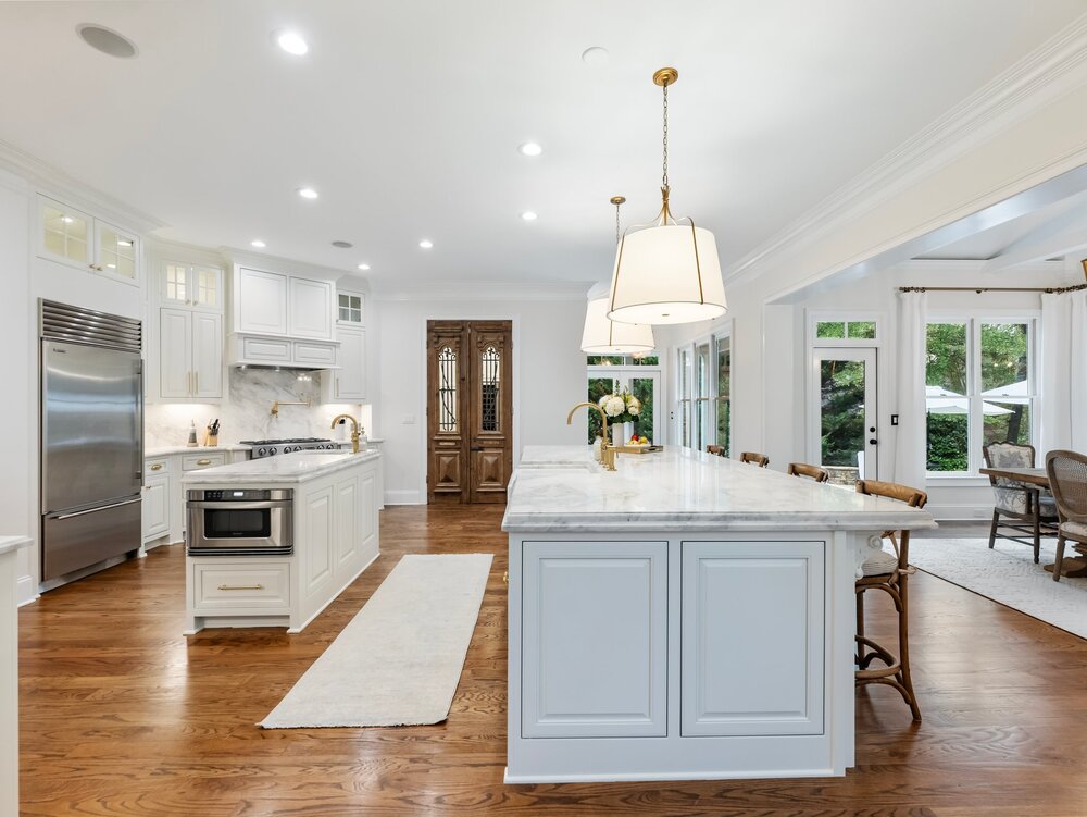 Spacious open-plan kitchen with large white marble-top breakfast island and hanging ceiling lamps and wood seats, another smaller work island, stainless steel refrigerator