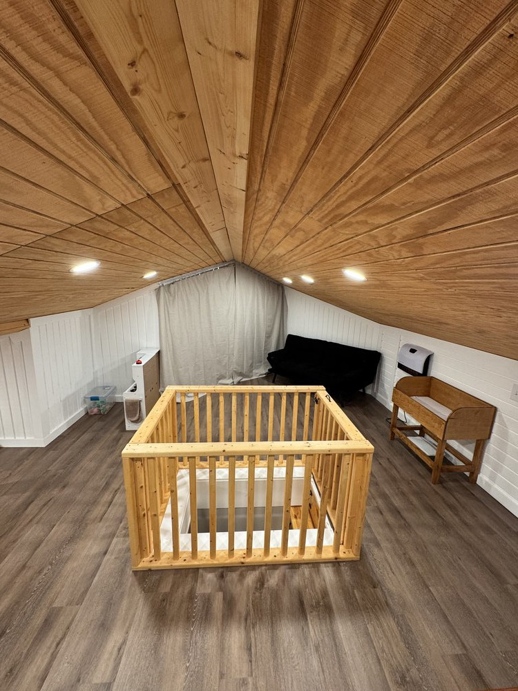 completed loft remodel with beautiful wood panel ceiling and recessed lighting, lovely laminate flooring, and furniture including a sofa, baby changing table, a play area, and wood-rail gate around the stairway access to prevent accidental falls