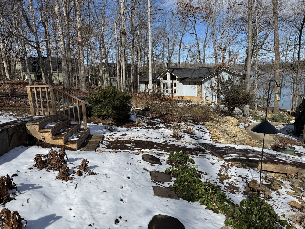 pathway to lake house showing old wood stairs with one-sided railing and four treads leading from upper ground area down to snow-covered lower ground