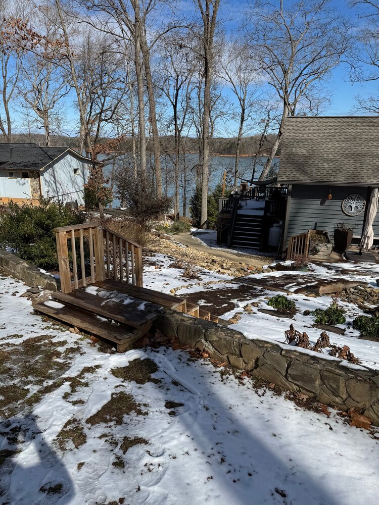 snow covered pathway to lake house with old wood stairs with four treads and one-sided railing leading from upper ground area down to a wood bridge coveering a creek bed