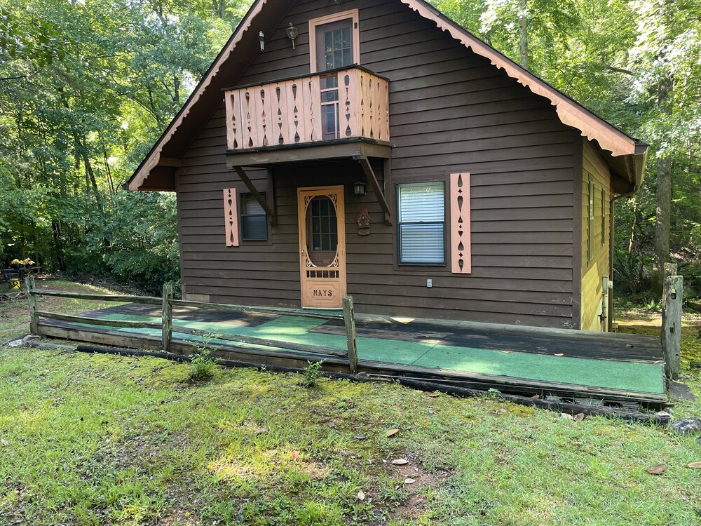 Before-photo of a brown wooden cottage-style cabin with a run-down deck, showing dark, rotten boards.